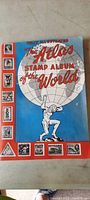 Blue front cover of The Atlas Stamp Album of the World showing illustrated globe with Atlas figure and surrounding small stamp images.