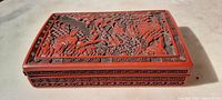 Top view of red lacquered rectangular box with intricate Asian-themed carvings on the lid.