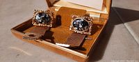 Close photo of pair of antique cuff links with ornate gold-tone setting and unique black and white speckled stones, shown in vintage brown velvet case