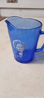 Blue glass pitcher with Shirley Temple white portrait, showing front side with spout, handle and base.