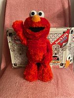 Red T.M.X. Tickle Me Elmo plush toy standing on pink cloth backdrop