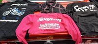 Four Snap-on Tools branded clothing items (black t-shirt with motorcycle graphic, plaid button-up, pink sweatshirt, black hoodie) arranged for lot identification.