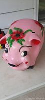 Front and side views of a large painted ceramic piggy bank shaped like a pig. The pig is pink with black hooves and facial features, decorated with red flowers and green leaves.