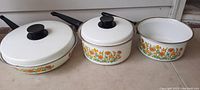 Front view of three metal cookware pans with white and sunflower design, two with lids and one without, arranged side-by-side.