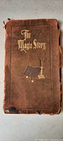 Front cover of the booklet showing leather material, gold embossed title 'The Magic Story' and decorative embossed imagery of book and candle holder.