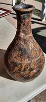 Close-up photo of the 12" tall clay pitcher/vase showing the neck, flared rim, and bulbous base with mixed black and orange coloration and horizontal incised markings.