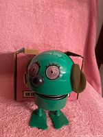Front view of Watergate Bug toy figure showing green metal body, spiral eyes, black nose and brown felt ears on pink fabric background