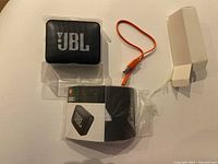 JBL GO 2 Bluetooth speaker with orange charging cable, plastic case, and packaging sleeve on a white surface.