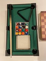 Miniature pool table showing green felt surface, two pool cues, triangle rack, and miniature pool balls; wooden chess set box to the right with checkered pattern on lid