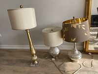 Three table lamps with metal bodies and fabric shades positioned against a wall. The lamps vary in height and style with metal bases in brass and silver tones and shades in cream, beige, and brown with leaf designs.