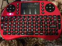 Front view of red mini wireless QWERTY keyboard with LED backlit keys, touchpad in center with gesture instructions, multimedia buttons on top left corner and directional pad on top right corner.