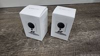Two WyzeCam V2 security cameras in original white packaging with printed product images, displayed on wood flooring.