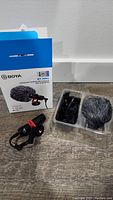 BOYA BY-MM1 microphone with box, furry windscreen, cable and mounting clip visible on wooden surface.