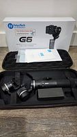 FeiyuTech G6 gimbal with original box, manuals, and carrying case on wooden floor surface.
