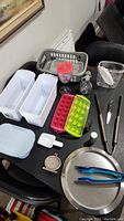 Photo showing various kitchen items including two white rectangular containers, pink and green ice cube trays, ice scoop, glass decanter, metal container, utensils and small baskets on black table.
