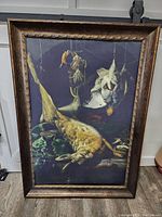 Front view of large framed canvas print showing a still life hunting scene with a hare and birds hanging amid vegetables, dark wood frame with textured border.