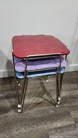 Stacked view of three stools with red, purple, and blue padded square seats and chrome metal legs.