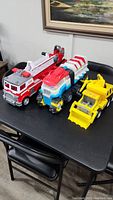 Three Paw Patrol toys arranged on a table showing firetruck, dinosaur vehicle, and construction vehicle