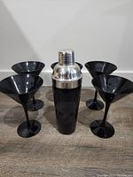 Full view of black cocktail shaker with silver top and four matching black martini glasses placed on wooden surface.