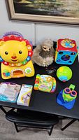 Display of all small child toys and safety items including lion push toy, sloth plushie, musical book, microscope, electronic toys and safety locks on table.