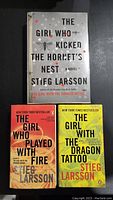 Three books of The Girl With The Dragon Tattoo trilogy by Stieg Larsson. The top book is a hardcover with gray cover titled 'The Girl Who Kicked the Hornet's Nest'. The bottom left is paperback 'The Girl Who Played With Fire' with an orange-yellow cover. The bottom right is paperback 'The Girl With The Dragon Tattoo' with yellow cover.