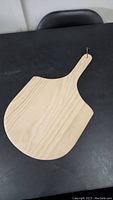 Wooden pizza peel showing the full blade and handle on a black surface, nail through the hanging hole