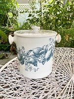 Front view of white ceramic chamber pot with lemon and foliage transfer pattern, two wide side handles, and covered lid with knob handle, placed on outdoor white woven table.