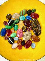 Thirty-five assorted vintage cabochons displayed on a yellow dish showing variety in shapes, colors, and sizes for sale.