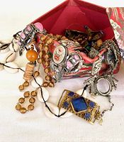 Jewelry pieces spilling from the red Asian fabric box showing pink glass-enamel metal segments, wooden beads, cowrie shells, and ornate pendants.