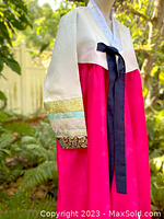 Side view of white cropped jacket with multicolored metallic brocade bands on the sleeves and navy tie closure over magenta skirt.