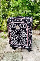 Full front view of upright spinner luggage with black and white damask pattern and pink piping, standing on outdoor stone surface with green foliage background.