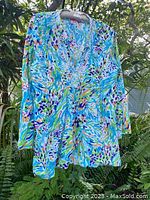 Front view of Lilly Pulitzer Sea Soiree tunic top hanging on a hanger outdoors with green foliage in background. Shows bright multi-color beaded embellishment and pattern.