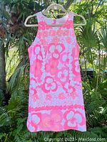 Front view of the sleeveless neon pink and orange floral cotton shift dress on hanger outdoors with green foliage.