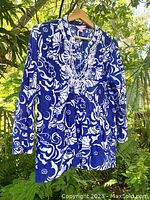 Front view of Lilly Pulitzer blue and white floral tunic top with white beaded neckline embellishment, hung on wooden hanger among green plants.