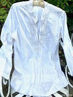 Front view of white cotton pintuck tunic top with beaded embellishment on collar and chest, displayed hanging outdoors against greenery.