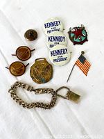 Top view of assorted vintage Americana pins, Kennedy for President buttons, Russian medal pin, and silver-tone bracelet with flask charm all displayed on white cloth background.