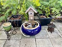 Full view of all garden and outdoor items including hose pot with hose and nozzles, birdhouse, two black planters, wrought iron plant stand with ceramic pot, and metal pagoda candle holder on paving stones with plants in background.