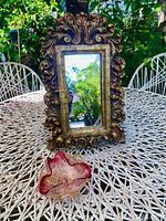 Baroque style gold ornate framed rectangular mirror and small pink and gold fleck Murano glass dish displayed on metal table outdoors.