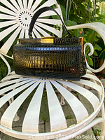 Black leather reptile textured handbag with brass metal closure sitting on white metal chair outdoors.