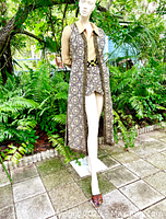 Full view of mannequin wearing the gold lame hotpants romper with the long brocade maxi vest outdoors surrounded by green plants.