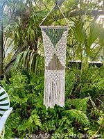 Macrame wall hanging shown suspended outdoors amidst plants, highlighting overall length and texture.