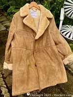 Full length front view of the light brown suede shearling lined coat hanging outdoors on a hanger.