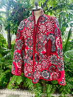 Front view of the heavy woven red, black, and white tapestry cape on mannequin outside with greenery background. Shows the floral pattern and front closure.