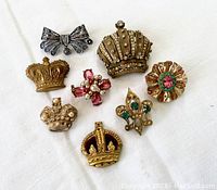 Seven assorted vintage brooches and pins including crowns, a bow, a fleur-de-lis, and a cross, all laid out on white fabric.
