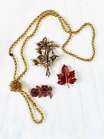 Photo showing full long amber-colored glass bead necklace with three different brooches arranged; flower brooch, maple leaf brooch with rhinestones, and pink rhinestone flower pin.