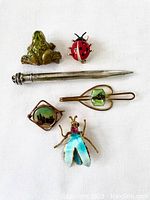 Group photo of six vintage jewelry items: silver-filled pencil pendant, green frog pin, red ladybug pin, European souvenir pin, enamel flying bug pin, and horse tie clasp.