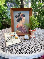 Full lot shown on outdoor table with greenery background, including framed painting, head vase, clock, book, and glasses.