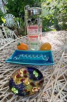 Vintage 1950s cocktail shaker with chrome top and colorful printed cocktail recipes, turquoise ceramic ashtray with ridge pattern, two bright orange ceramic salt and pepper shakers, and a small floral-patterned enamel dish on metal base shown together on outdoor table