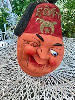 Close-up front and angled view of the vintage Shriners Egypt mascot face showing the painted plaster or chalkware material, red fez with gold detailing, and detailed facial features with winking expression.