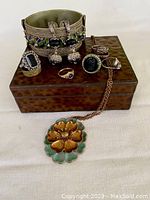 Vintage handmade slag glass trinket box with jewelry on top including a bangle, earrings, necklace with floral pendant, and several rings.